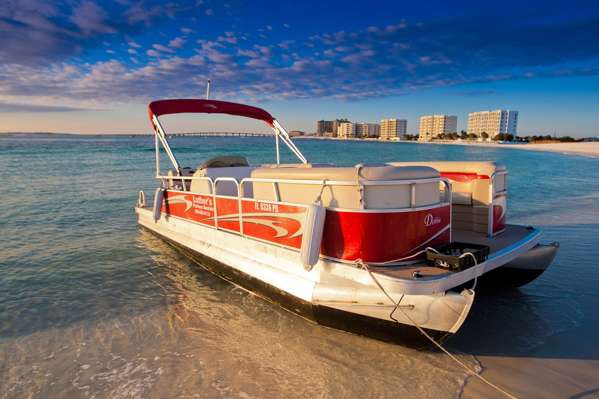 A Luther's pontoon boat beached on white sand in clear water at sunset, Destin condos behind